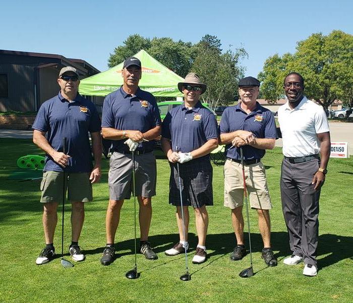 A group photo of 5 men with their golf tees in front of a bright green tent.