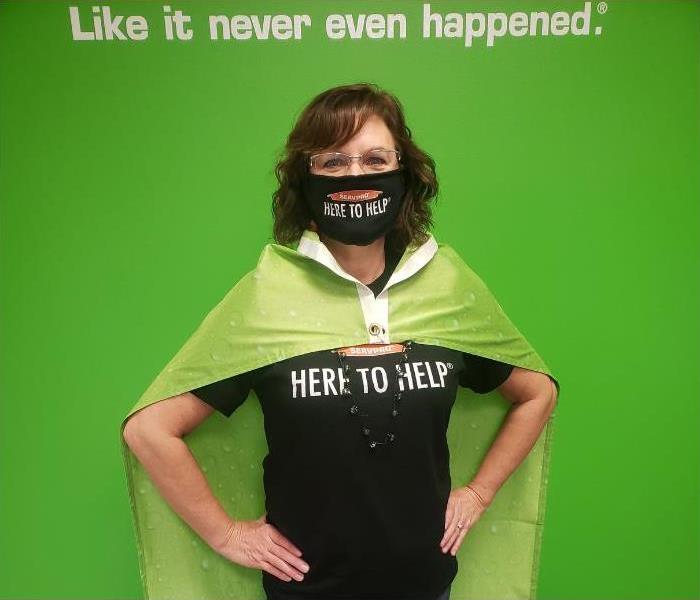 A woman wearing a black mask, a shirt that says here to help and a green cape.