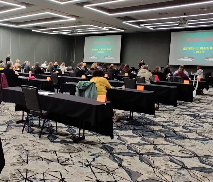 A event center with several tables spaced apart, and one person per table listening to a presenter.